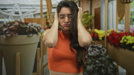 Young hispanic woman covers ears at crowded flower shop building aisle filled with colorful blooms; distress.