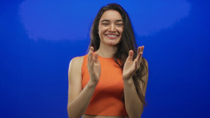 Young hispanic woman clapping hands in blue studio wearing orange top, smiling broadly; celebration.