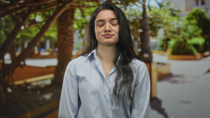 Young hispanic woman with long dark hair in striped shirt winks eye on leafy sunlit street corner;...