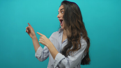 Young hispanic woman with wide eyes and open mouth pointing finger in turquoise studio space;...
