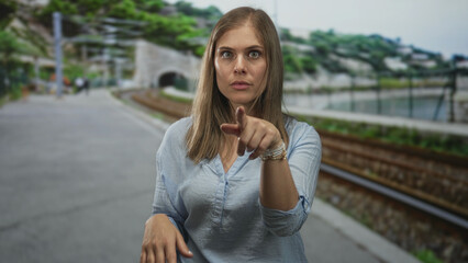 Fototapeta premium Woman with hands to face and pointing finger on street near railway tracks, open mouth gesture and raised palms; surprise doubt and questioning.