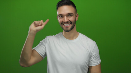 Fototapeta premium Young hispanic man smiling and pointing up in front of a vibrant green background, wearing a white t-shirt with a joyful expression, captured in a studio setting