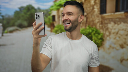 Young hispanic man wearing white shirt having a video call with a smartphone outdoors on a cobblestone street in an old town city setting with greenery and rustic buildings.