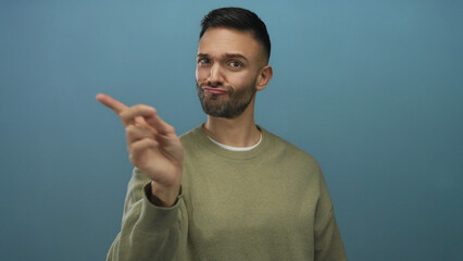 Young man gesturing with finger against blue background, wearing casual sweater, expressing serious...