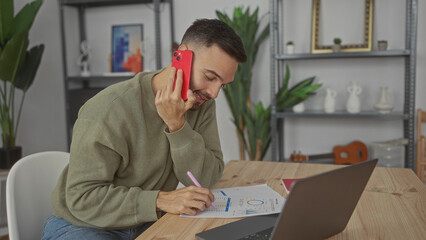 Young man talking on phone while writing notes in living room home office setting with laptop, highlighting casual work environment.