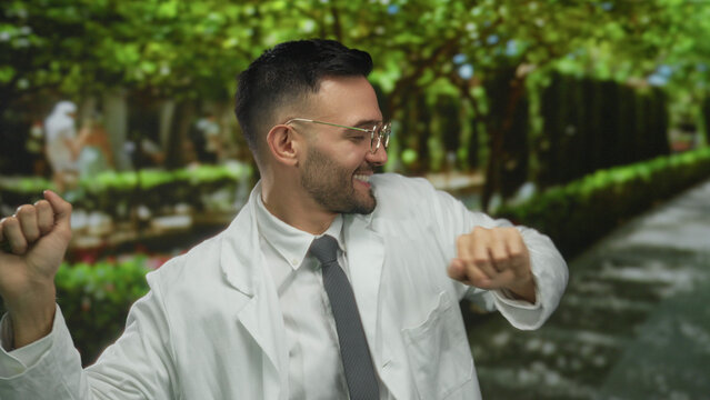 Young hispanic man in a white coat joyfully dancing in a green outdoor park setting, exuding happiness and energy with trees and paths in the background.