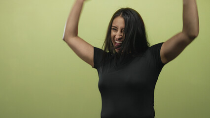 Woman cheering with open mouth and raised arms, young, wearing black top in green studio;...