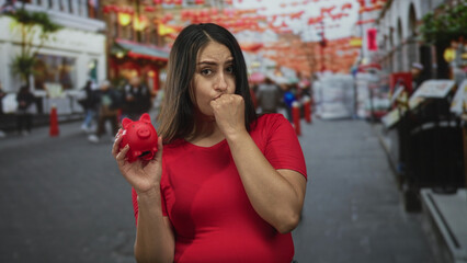 Woman in red shirt holding a red piggybank, biting her nails and clutching the coin box on a busy city street lined with storefronts and lanterns; financial worry.