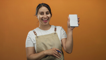 Woman in apron holding a notepad and pen, smiling and showing her hand in an orange studio;...