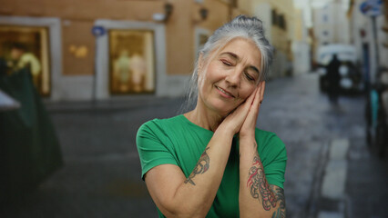Senior woman with grey hair smiling and making a sleeping gesture with hands on a street, showcasing her tattoos in an outdoor setting.