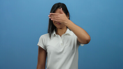 Young latin woman in white shirt covering her face with hand in stop gesture against blue...