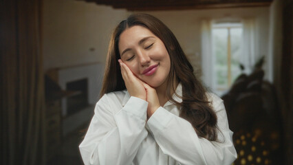Young hispanic woman with eyes closed and hands pressed to her cheek in a sunlit studio room mimicking sleep; serenity.