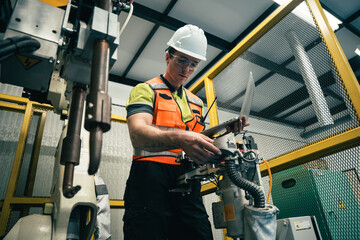 Engineer in safety gear using laptop to inspect and control industrial robot in modern factory,...
