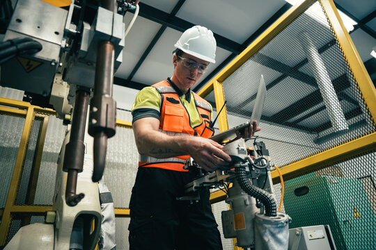 Engineer in safety gear using laptop to inspect and control industrial robot in modern factory, concept of automation, robotics, smart industry, and advanced technology. AI machine learning