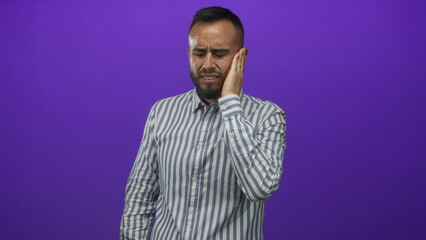 Young hispanic man in a striped shirt holding hand to ear and wincing in a purple studio with toothache; pain.