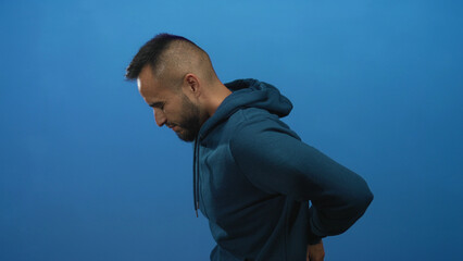 Man holding lower back while wincing and clasping waist, wearing hoodie in blue studio for...