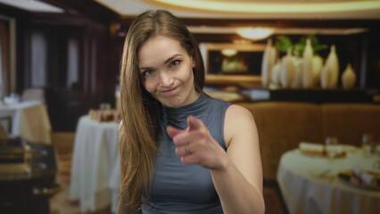 Woman beckons with index finger at viewer amid blurred tables and decor in restaurant interior; invitation anticipation.