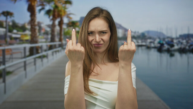 Woman holds up two middle fingers at a port dock under bright daylight, scowling intensely at the lens; defiance.