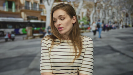 Fototapeta premium Woman in striped top with eyes closed covers mouth yawning on crowded city street lined with trees and passing bus; fatigue.