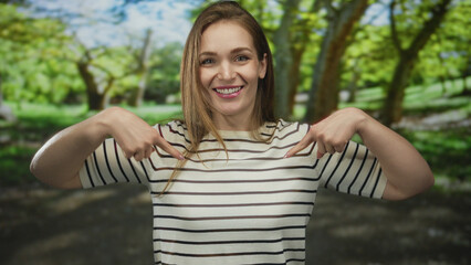 Young caucasian woman in white striped shirt points finger to chest in forest amid green trees;...