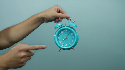 Man's hand holding a turquoise alarm clock against a blue background, with a pointing gesture...