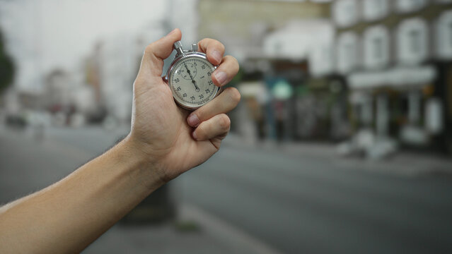 Hand holding a stopwatch in an urban street setting with blurred town buildings, symbolizing time management and precision by a man in an outdoor city environment.
