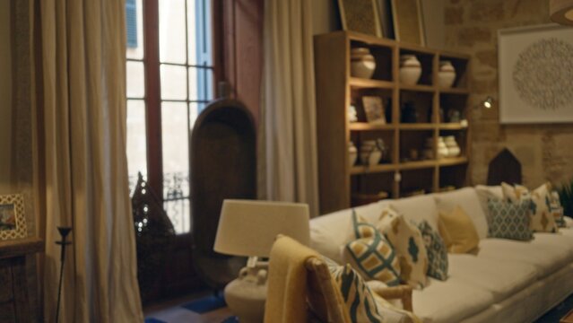 Cozy living room with soft focus decor, bokeh effect on elegant furniture and patterned cushions, shelves displaying vases, framed by large windows and soft curtains.