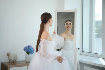 Naklejka premium Dreams come true. Woman wearing beautiful wedding dress looking at herself in mirror indoors