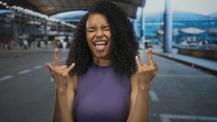 Woman making rock gesture while smiling outdoors at busy airport terminal with modern architecture...
