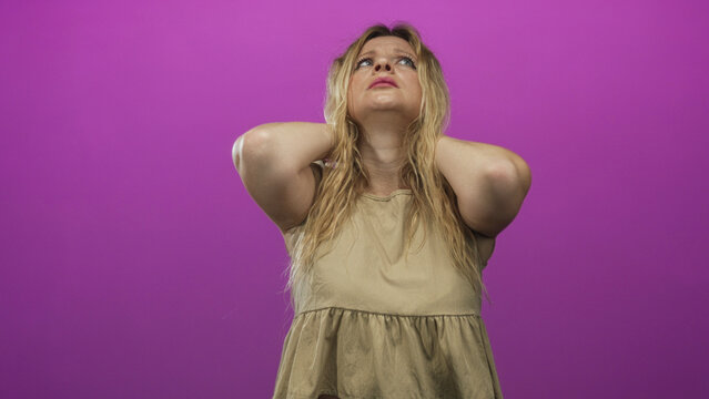 Young blonde woman holding head with hands, bare arms visible, tense worried facial expression in purple studio set; anxiety concern unease.