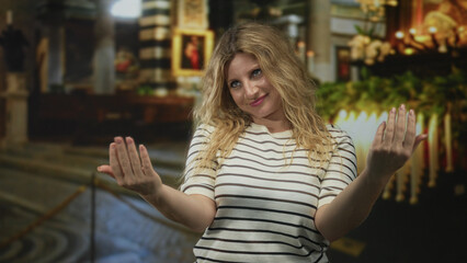 Young blonde woman in striped shirt beckons with both hands amid church aisle and votive candles; welcoming invitation calm.