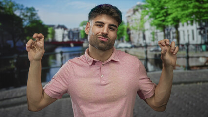 Naklejka premium Young hispanic man in pink polo shirt showing playful smirk with hands making quote gesture on a street; sarcasm.