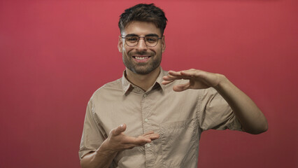Man with hands framing imaginary object in a red studio wearing round glasses and beige shirt; confidence.