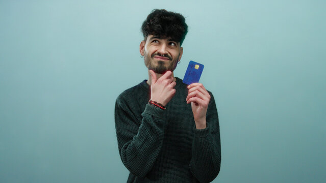 Man holding a blue credit card against a blue background looks thoughtful with a slight smile suggesting decision making or financial consideration.