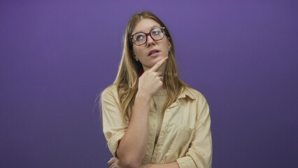Young blonde woman wearing glasses rests hand on chin gesture in studio with purple backdrop; thoughtfulness reflection contemplation pondering.