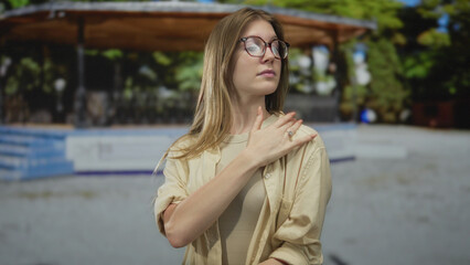 Woman flips long blonde hair beside wooden pavilion in lush green park under soft sunlight;...