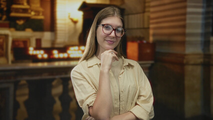Woman wearing glasses with hand on chin while standing before a candlelit altar in church; serenity.