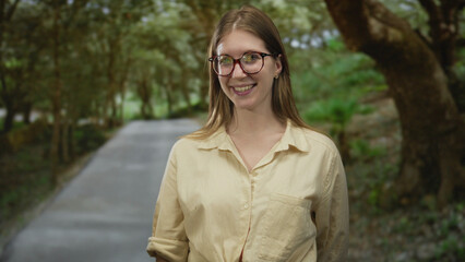 Blonde woman wearing glasses smiles while standing on forest path under filtered green sunlight; serenity.