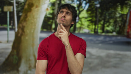 Man in red shirt standing beside tree with hand on chin for thinking in park with green foliage;...