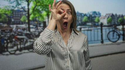 Fototapeta premium Woman gestures ok sign with hand along sunlit canal embankment on a lively bustling city street; approval.