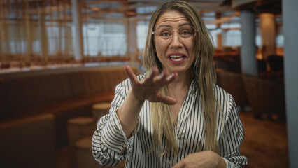 Woman with long blonde hair wearing round glasses and striped shirt with hand to lips gesture in...
