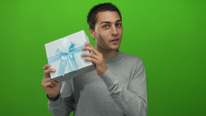 Young man examining a gift box in front of an isolated green wall, wearing a gray shirt, conveying...