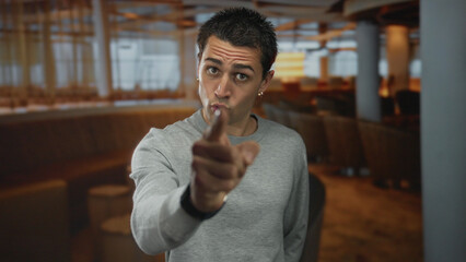 Young man indoors pointing finger at restaurant wearing gray sweater and earrings with serious...