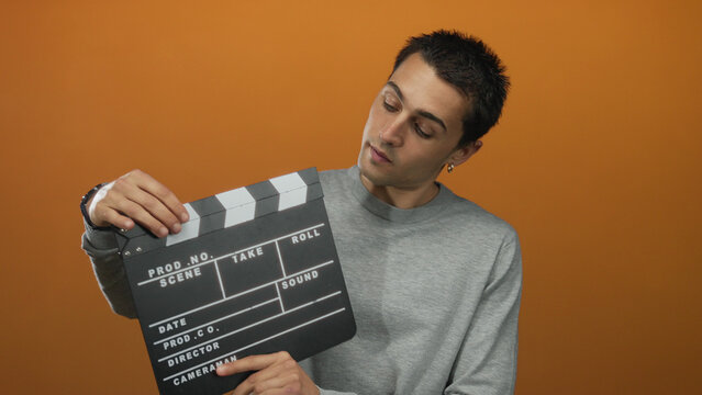Young hispanic man in gray sweater holding clapperboard against isolated orange background, capturing creative filmmaking and production concepts.