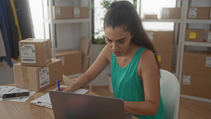 Woman writing on package label beside stacked cardboard parcels at wooden desk in building; concentration.