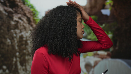 African american woman holds smartphone in park surrounded by green foliage and touches forehead;...