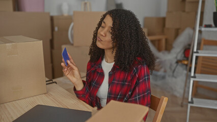 Woman in a plaid shirt holds a creditcard and gazes at it among cardboard boxes inside a building; financial uncertainty.
