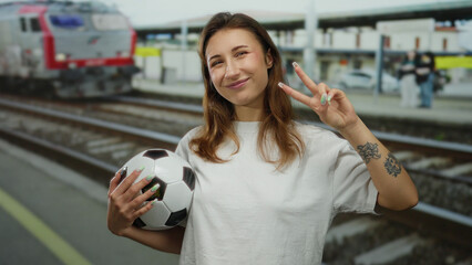 Woman smiling at train station holding a soccer ball showcasing peace sign with a tattoo visible against a backdrop of rail tracks and a passing train.