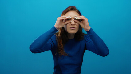 Woman peering through fingers on isolated blue background suggests curiosity, with a caucasian...