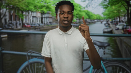 Fototapeta premium Man points finger upward at canal edge in city street framed by leafy trees; curiosity and discovery.